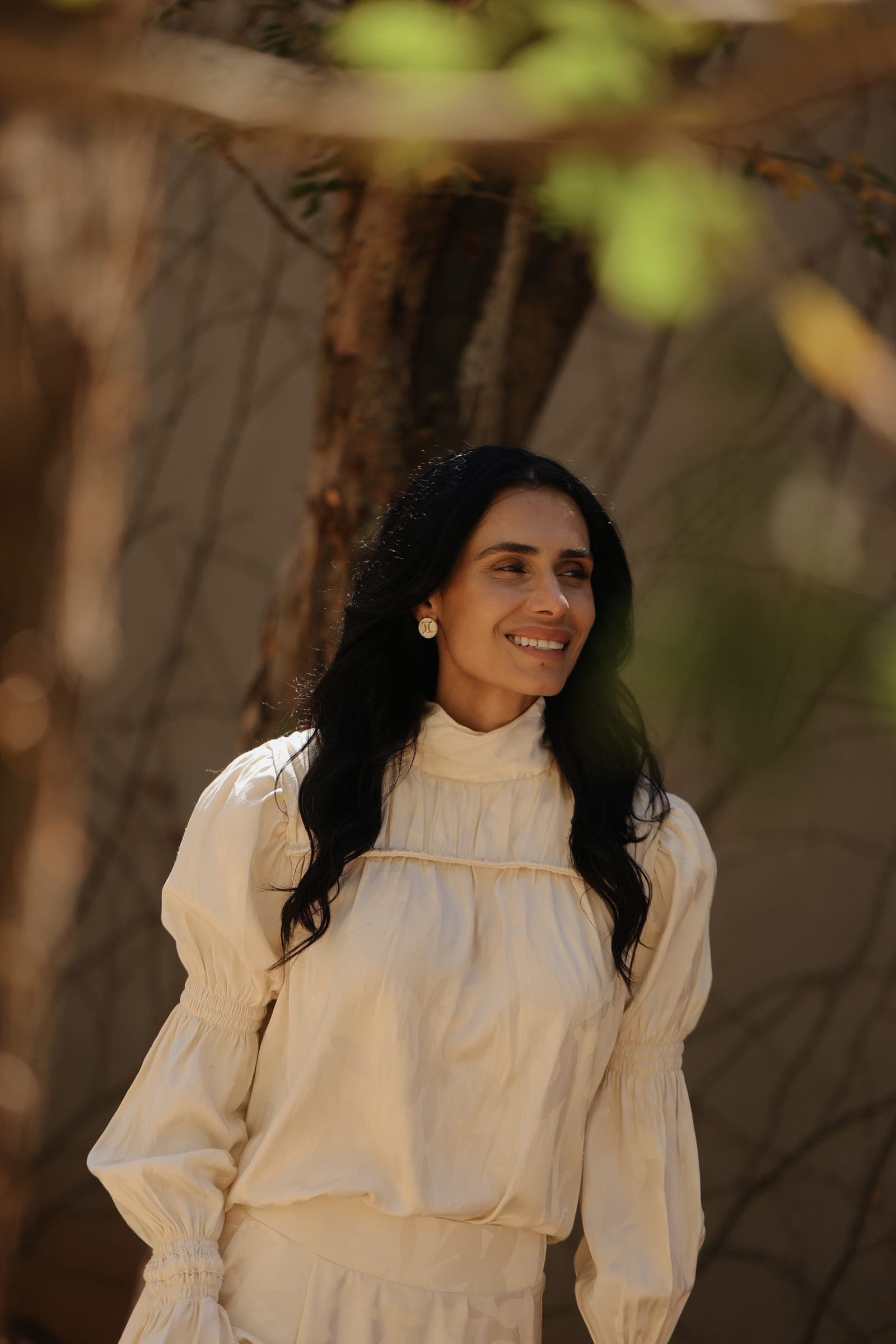 Mulher de vestido cru junto a uma oliveira, olhar sereno